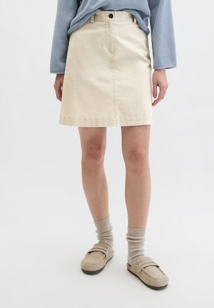 Frau trägt einen beigen knielangen Rock, hellblauen Pullover, beigen Socken und beige Slip-On-Schuhe vor weißem Hintergrund.