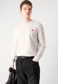Pull à manches longues crème clair avec des poignets côtelés, arborant un petit patch logo rouge sur la poitrine, associé à un jean noir fuselé et une veste en cuir noire.