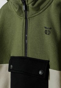 Veste color-block verte, blanche et noire avec fermeture éclair à l'avant, logo sur la poitrine indiquant "Ninety7" au-dessus de deux icônes de globe, et rabat de poche à bouton-pression.