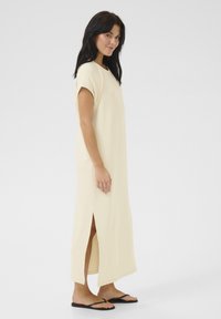 Rochie maxi galben deschis, cu mâneci scurte, slituri laterale și textură de țesătură netedă, asortată cu sandale negre. Design simplu și modern.
