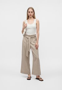 Mujer joven con pantalones beige de talle alto y pierna amplia y top blanco sin mangas, sosteniendo un bolso blanco tejido al hombro, y calzando chanclas negras.