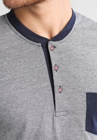 Polo à manches courtes gris rayé avec un col bleu marine, patte de boutonnage à quatre boutons et une poche sombre, présentant des accents décoratifs rouges et noirs.