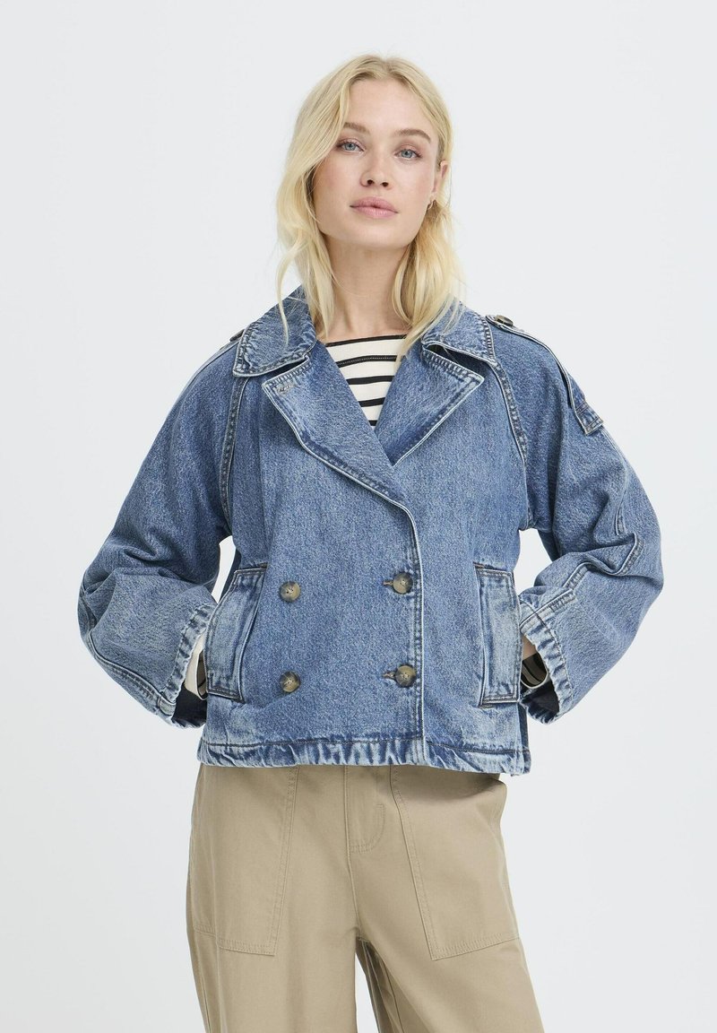 Veste en denim à double boutonnage avec un col structuré, de grandes poches et un délavage distinctif, portée avec une chemise rayée en dessous.