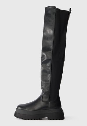 Botas pretas acima do joelho com um acabamento em couro liso, sola plana e sola de borracha robusta. Apresenta um fecho lateral e painéis texturizados.