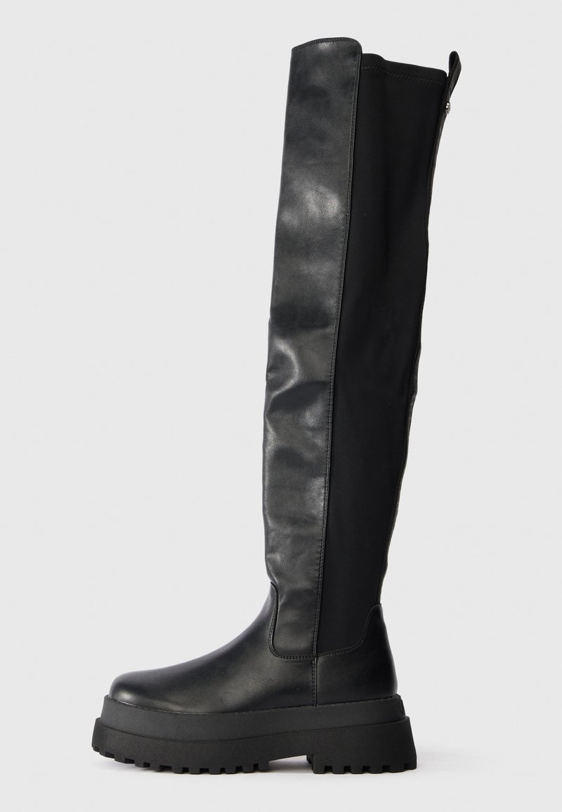 Botas pretas acima do joelho com um acabamento em couro liso, sola plana e sola de borracha robusta. Apresenta um fecho lateral e painéis texturizados.