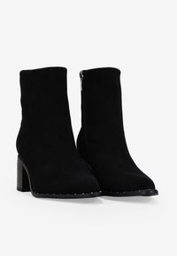 Botines de ante negros hasta el tobillo con puntera puntiaguda, tacón cuadrado, cremallera lateral y detalles de tachuelas a lo largo de la suela. Textura suave y diseño elegante.