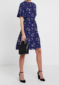 Marinblommig klänning med korta ärmar, insvängd midja och utsvängd kjol; i kombination med en liten svart handväska och svarta högklackade skor.