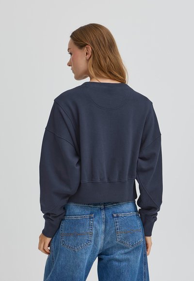 Tmavomodrý skrátený teplák s padnutými ramenami, textúrovaná látka a rebrovaný lem. Skombinovaný s svetlomodrými džínami s vreckami na zadnej strane.