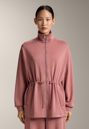 Frau trägt eine staubrosa Jacke mit Reißverschluss, Kordelzug in der Taille und langen Ärmeln, steht vor einem neutralen Hintergrund.