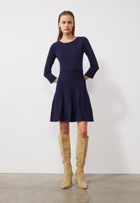 Robe en tricot bleu marine avec des manches trois-quarts, corsage ajusté et jupe évasée avec des plis. Associée à des bottes en suède beige jusqu'aux genoux.