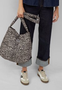 Persoon die een gepolsterde tote bag met luipaardprint vasthoudt, gekleed in donkere omgeslagen spijkerbroek en witte sneakers met zwarte en beige details.
