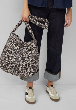Persona che tiene una borsa a tracolla imbottita con stampa leopardata, indossando jeans scuri con risvolto e sneakers bianche con dettagli neri e beige.