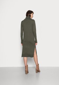 Robe en tricot vert olive avec un col roulé, des manches longues et des fentes sur les côtés. Elle a une silhouette ajustée, associée à des bottines en cuir beige.
