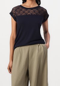 Marineblå t-shirt med blondeoverlayer, der har et geometrisk mønster, korte ærmer og en afslappet pasform, sammen med beige teksturerede bukser.