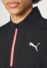 Giubbotto sportivo nero con colletto alto, dotato di una zip rosa e di un logo Puma bianco sul lato destro. Tessuto liscio e leggero.