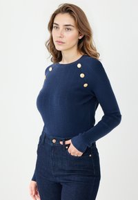 Navy niebieski sweter dzianinowy z pionowymi prążkami i złotymi guzikami na ramionach. Połączony z ciemnymi dżinsami w dopasowanej sylwetce.