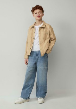Junge Person mit lockigem Haar steht vor einer einfarbigen Wand, trägt eine beige Jacke, ein weißes Hemd, eine patchworkartige weite Jeans und weiße Sneaker.