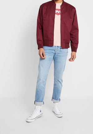 Veste bomber bordeaux avec un col côtelé, une fermeture éclair et des poches latérales, portée par-dessus un t-shirt rose pâle et associée à un jean bleu clair et des baskets blanches.