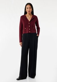 Burgunderfarbige, kurz geschnittene Strickjacke mit Reverskragen, goldenen Knöpfen und aufgesetzten Taschen, kombiniert mit hochtaillierten schwarzen Hosen.