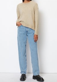 Pull en maille beige avec une coupe décontractée et un ourlet côtelé, associé à un jean en denim bleu clair. Des chaussures noires à semelle épaisse complètent la tenue.