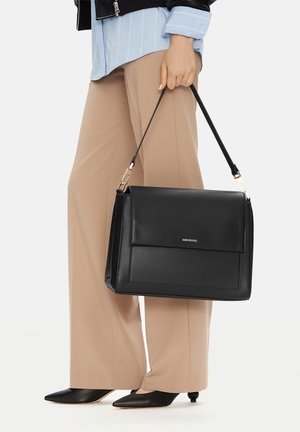 Mujer sosteniendo un bolso de cuero negro con herrajes dorados, vistiendo pantalones beige, zapatos negros puntiagudos, camisa azul y chaqueta negra.