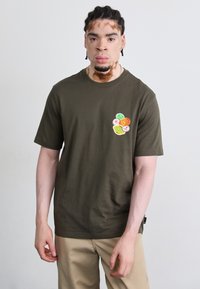 Olivengrøn t-shirt lavet af bomuld, med et farvet grafisk design bestående af cirkulære former på venstre brystområde. Korte ærmer, rund hals.