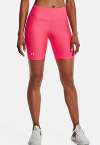 Sportovec oblečený v jasně růžových cyklistických šortkách Under Armour a bílých sportovních botách, stojící na bílém pozadí.