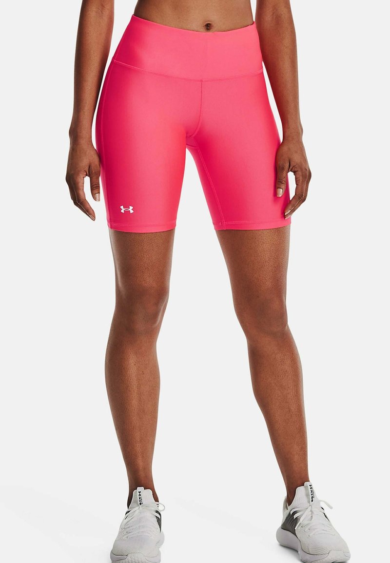 Sportovec oblečený v jasně růžových cyklistických šortkách Under Armour a bílých sportovních botách, stojící na bílém pozadí.