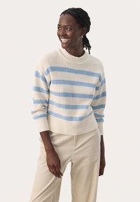 Pull rayé en crème avec des rayures bleues, présentant une texture côtelée et un col montant. Associé à un pantalon en velours côtelé beige, offrant une coupe décontractée.