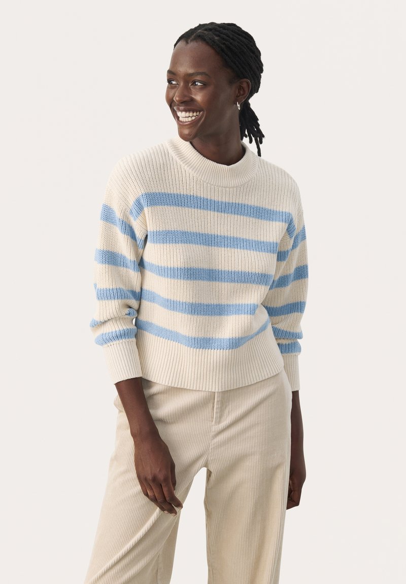 Pull rayé en crème avec des rayures bleues, présentant une texture côtelée et un col montant. Associé à un pantalon en velours côtelé beige, offrant une coupe décontractée.