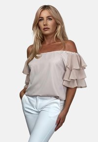 Bluzka odsłonięta na ramionach w jasnoróżowym materiale, z warstwowymi falbanami na rękawach. Połączona z białymi dopasowanymi spodniami. Gładka tekstura.