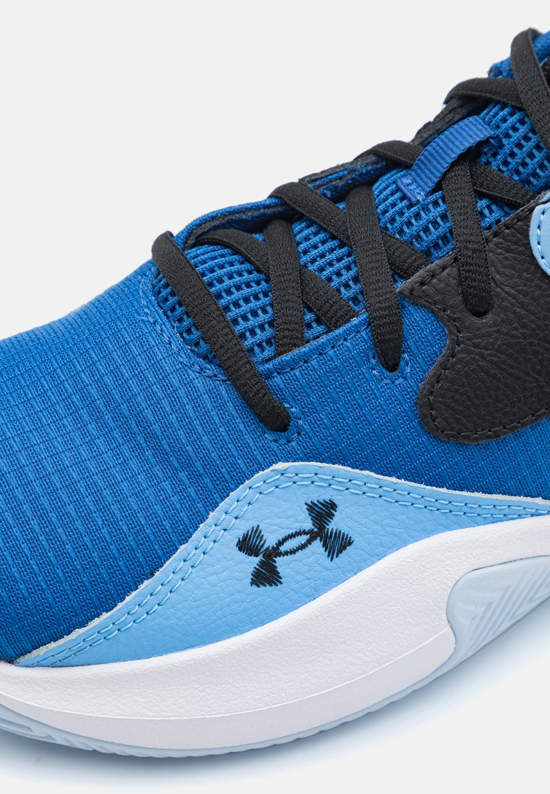 Gros plan sur une chaussure de sport bleue avec des lacets noirs, le logo Under Armour et une semelle blanche sur un fond blanc.