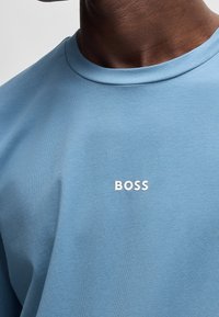 Světle modré bavlněné tričko s kulatým výstřihem, na hrudi se bílým logem "BOSS". Tkanina je hladká s mírnou texturou.