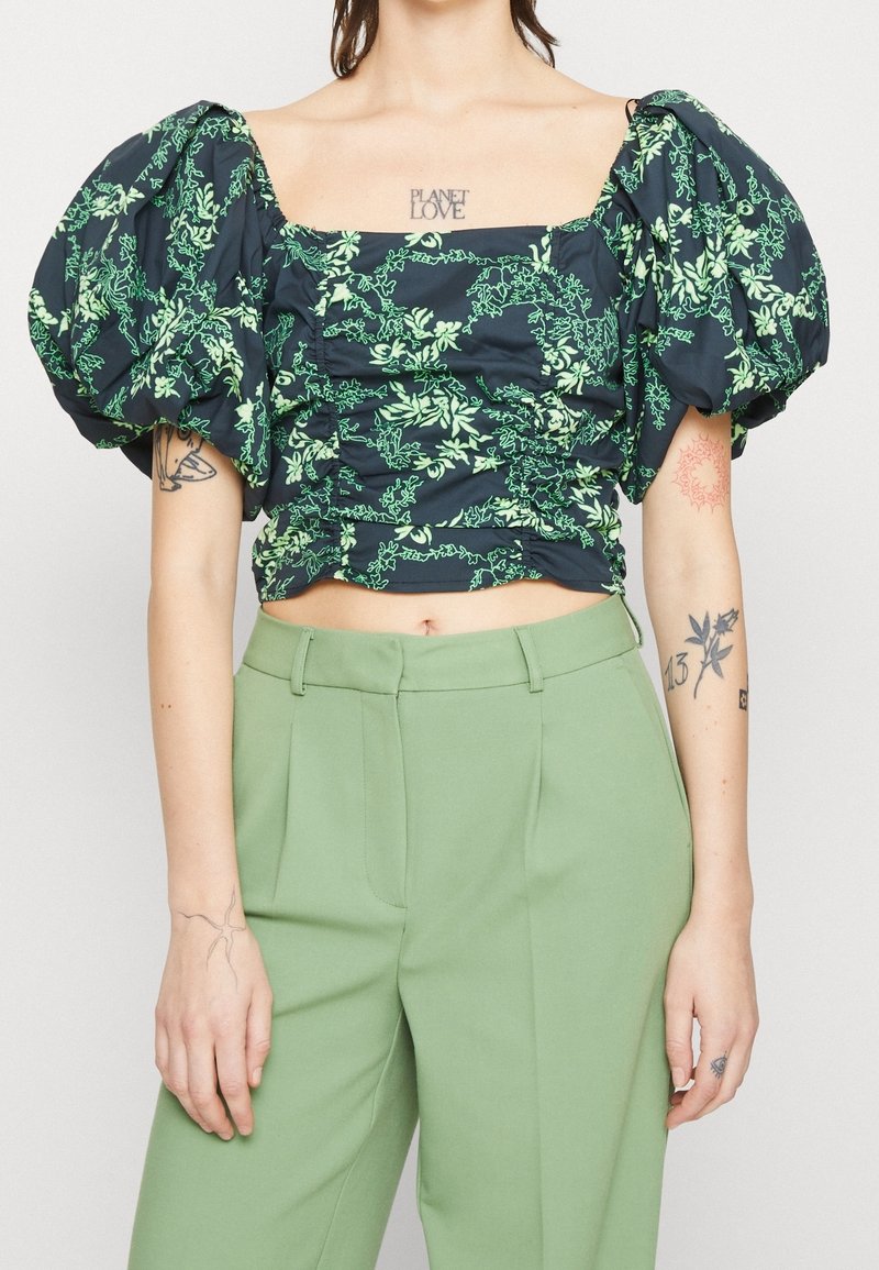 Blusa nera con maniche a palloncino e stampa floreale verde, texture arricciata e scollatura quadrata, abbinata a pantaloni su misura verde chiaro.