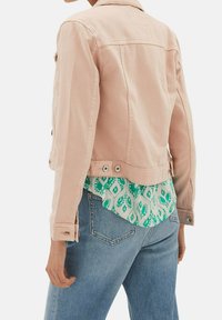 Jasnoróżowa denimowa kurtka z klasycznym kołnierzykiem, zapięciem na guziki i bocznymi kieszeniami; nałożona na wzorzystą zieloną i białą koszulę.