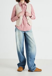 Gilet in corduroy marrone chiaro sopra una camicia a quadri rossa, abbinato a jeans a gamba larga blu sbiaditi e scarpe slip-on marroni. Sfondo neutro.