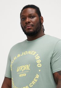 T-shirt in cotone verde chiaro con grafica circolare gialla contenente il nome del marchio e la data. Collo a giro e maniche corte, texture liscia.