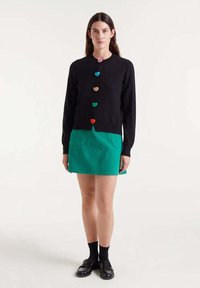 Schwarze Strickjacke mit herzförmigen Knöpfen in Rot, Pink, Grün und Orange, kombiniert mit einem grünen Cord-Minirock. Glatte Textur, taillierte Passform.