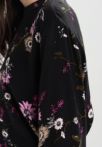Vêtement noir avec un motif floral comportant des fleurs roses, blanches et beiges. Coupe ample avec un col rond et des manches courbées.