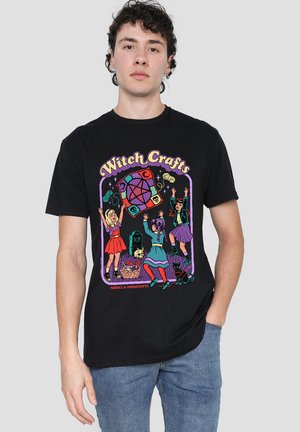 Junger Mann trägt ein schwarzes T-Shirt mit einem bunten Cartoon von drei Mädchen, die mit Garn und Hexerei-Utensilien spielen.