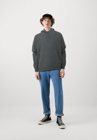 Šedá mikina s kapucí a přední kapsou, v kombinaci se světle modrými džíny a černými vysokými teniskami. Minimalistický design, pohodlný střih.