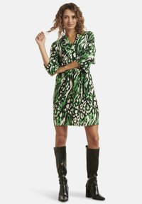 Grünes und schwarzes gemustertes Kleid mit Kragen und Dreiviertelärmeln, das eine taillierte Taille hat. Model trägt schwarze kniehohe Stiefel.