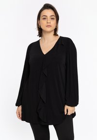 Blouse noire à manches longues avec un col en V, arborant un devant froncé et un tissu texturé. Le design est ample et arrive aux hanches.