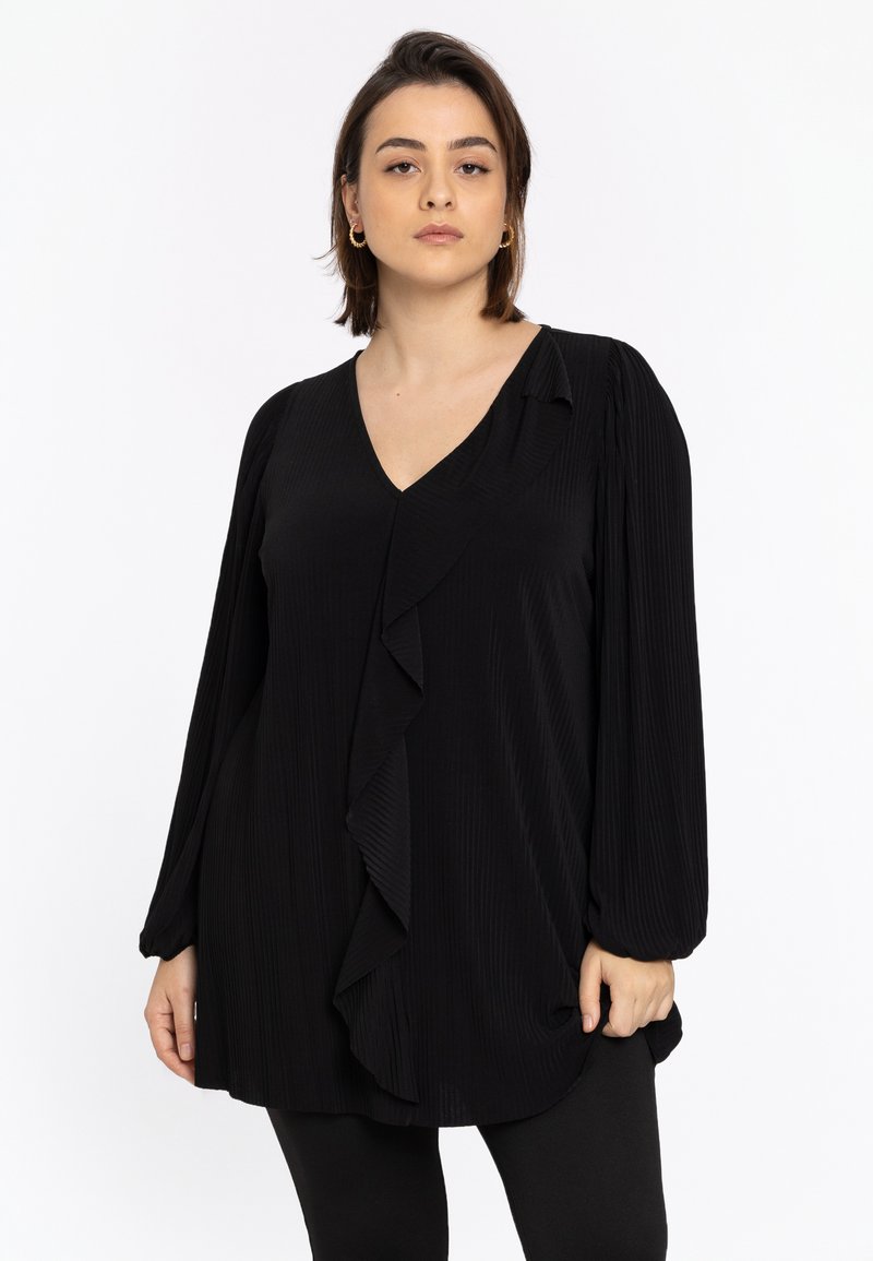 Blouse noire à manches longues avec un col en V, arborant un devant froncé et un tissu texturé. Le design est ample et arrive aux hanches.
