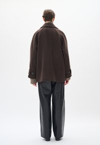 Abrigo de lana marrón con un cuello estructurado, visto desde atrás, combinado con pantalones de cuero negros de pierna ancha, que muestran una silueta relajada.