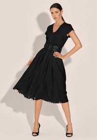 Schwarzes Lochstickkleid mit kurzen Ärmeln, V-Ausschnitt, tailliertem Bund und ausgestellt Rock. Detaillierte Lochmuster und gewellter Saum. Schwarzer Gürtel enthalten.