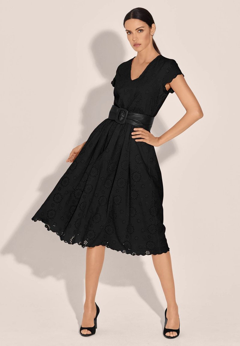 Schwarzes Lochstickkleid mit kurzen Ärmeln, V-Ausschnitt, tailliertem Bund und ausgestellt Rock. Detaillierte Lochmuster und gewellter Saum. Schwarzer Gürtel enthalten.
