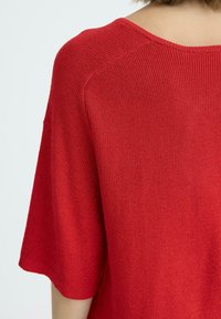 Crvena pletena majica s kratkim rukavima, s širokim izrezom i rebrastom teksturom. Tkanina izgleda mekano i blago rastezljivo.
