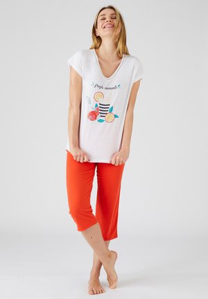 Glunderende vrouw staat op blote voeten in wit V-hals T-shirt met print van citrusdrank en oranje cropped broek tegen een effen lichte achtergrond.