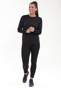 Schwarzes langärmliges Sporttop mit Rundhalsausschnitt, kombiniert mit schwarzen enganliegenden Leggings. Getragen mit schwarzen Turnschuhen, aus glattem Material und mit einem eleganten Design.